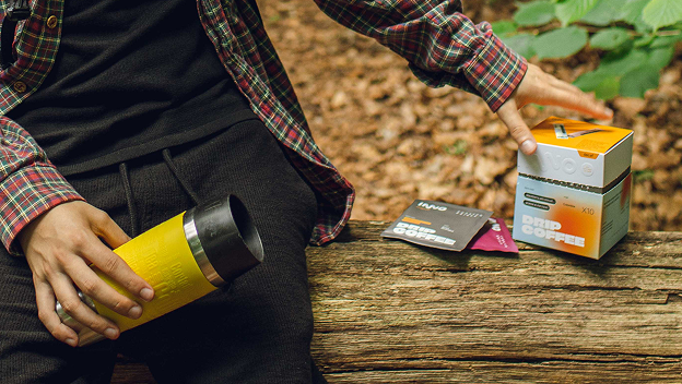Person outdoors with a yellow water thermos and a box of specialty drip coffee on a wooden surface.