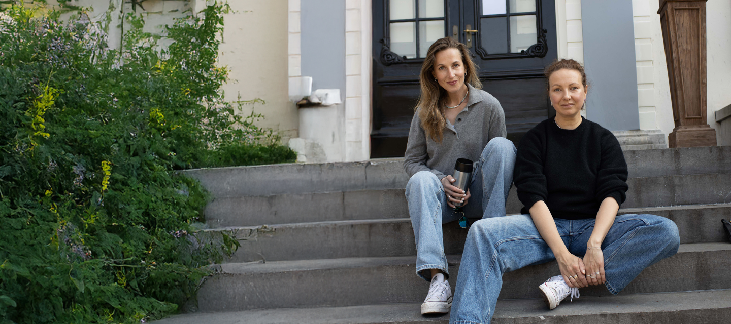 Two women - bussines founders sitting on steps outside a house with plants and a door in the background.