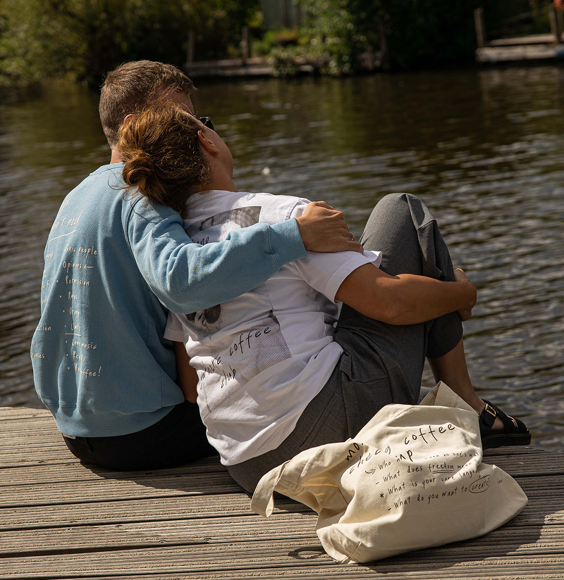 Two people sitting on a dock by a body of water, with a 'specialty coffee' bag in front of them.